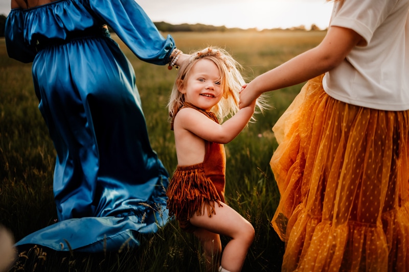 Family Photographer, A toddler girl is holding the hands of two woman family members as they walk through a grassy field